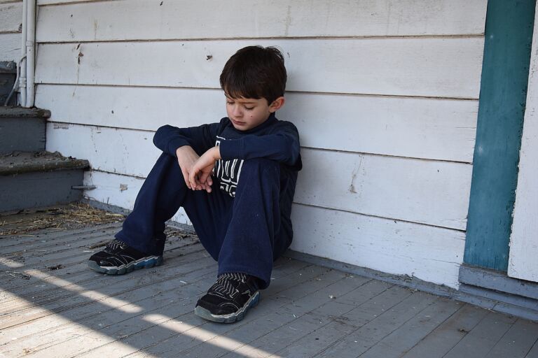 Male child around 6-10 years of age sitting down out on a porch. His head is down looking upset