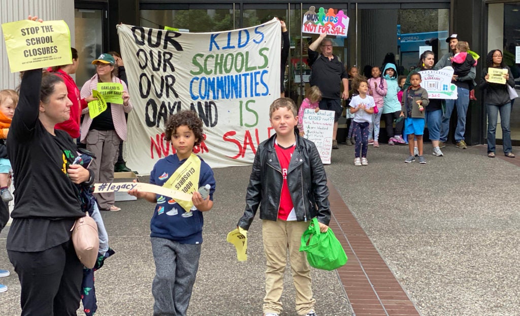 An elementary school teacher holds a protest sign alongside two elementary-aged kids